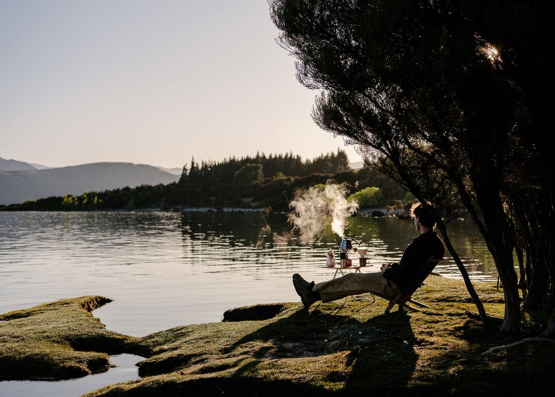 Traveler sits with hot drink on side of scenic lake