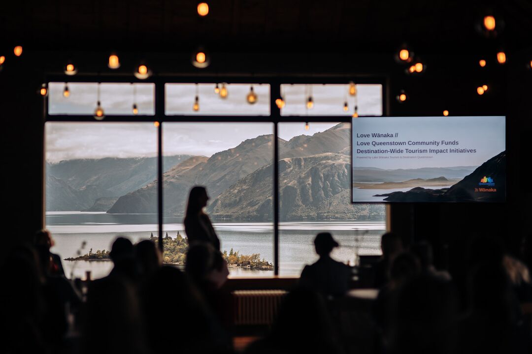 Room interior with people silhouetted against large windows. View of lake and mountains outside. String lights above, information screen visible