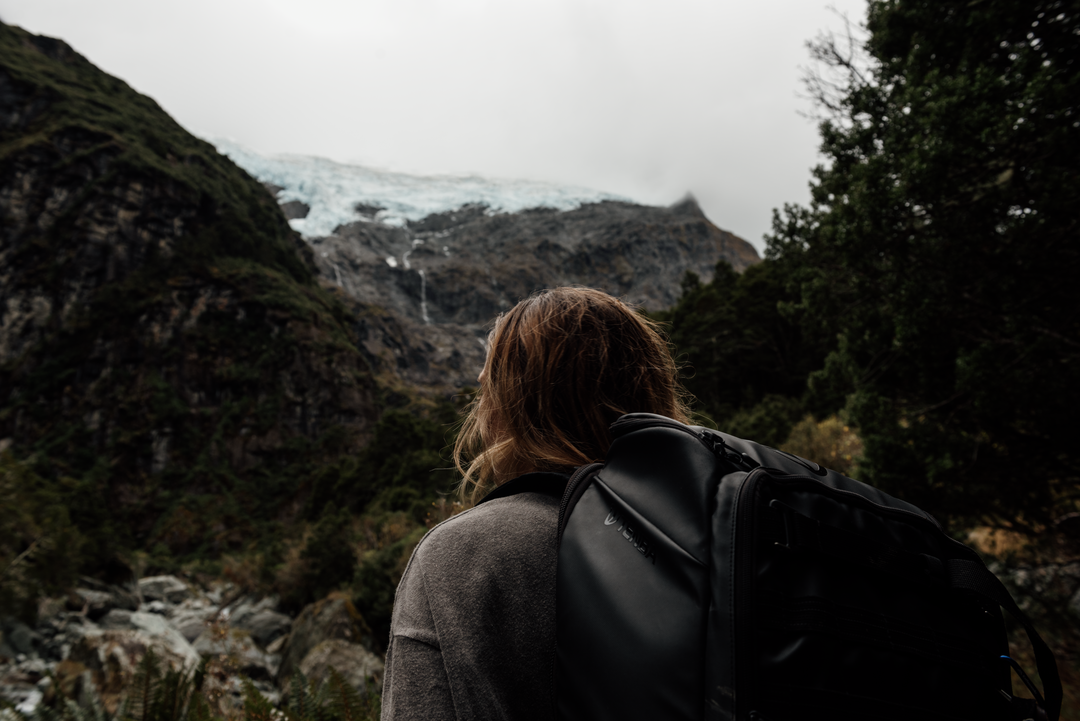 Hiker overlooking Rob Roy Glacier, Mount Aspiring National Park, New Zealand, with backpack facing rugged mountains and distant blue ice.