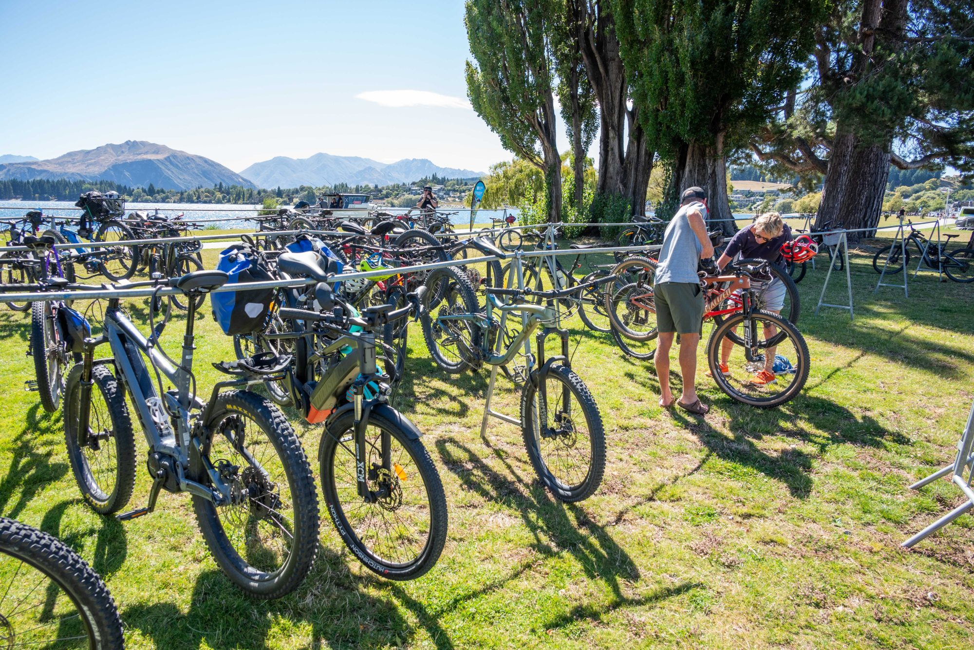A bike parking area by Lake Wānaka, filled with mountain bikes on temporary bike racks. Two people are tending to a bike in the background. The scene shows Lake Wānaka and mountains in the distance, with large poplar trees providing shade