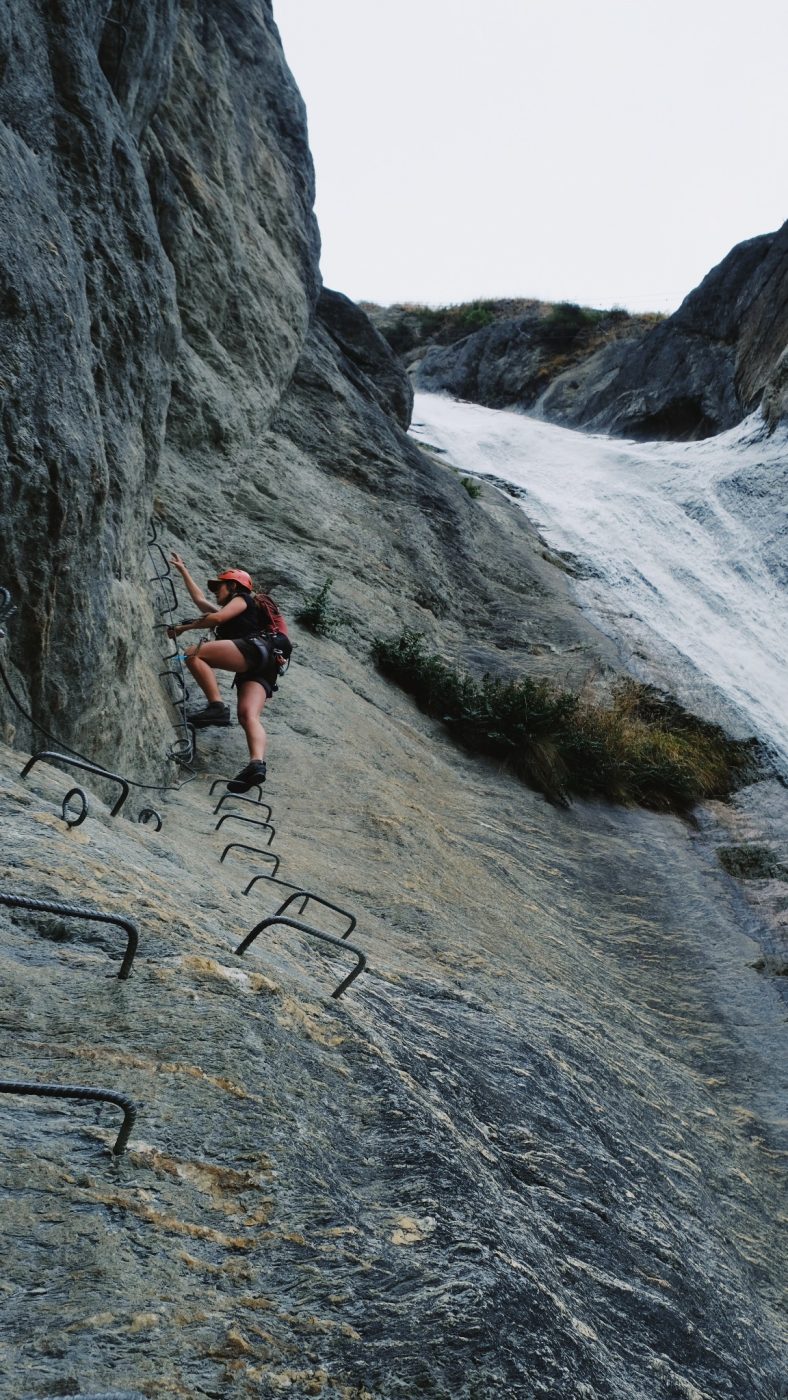 a female climbing up a runged ladder on a rock face with a waterfall beside them at Wildwire at Wānaka New Zealand