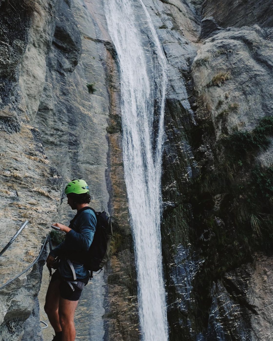 A climber in a green helmet standing next to a rockface with a waterfall in the background at Wildwire Wānaka New Zealand