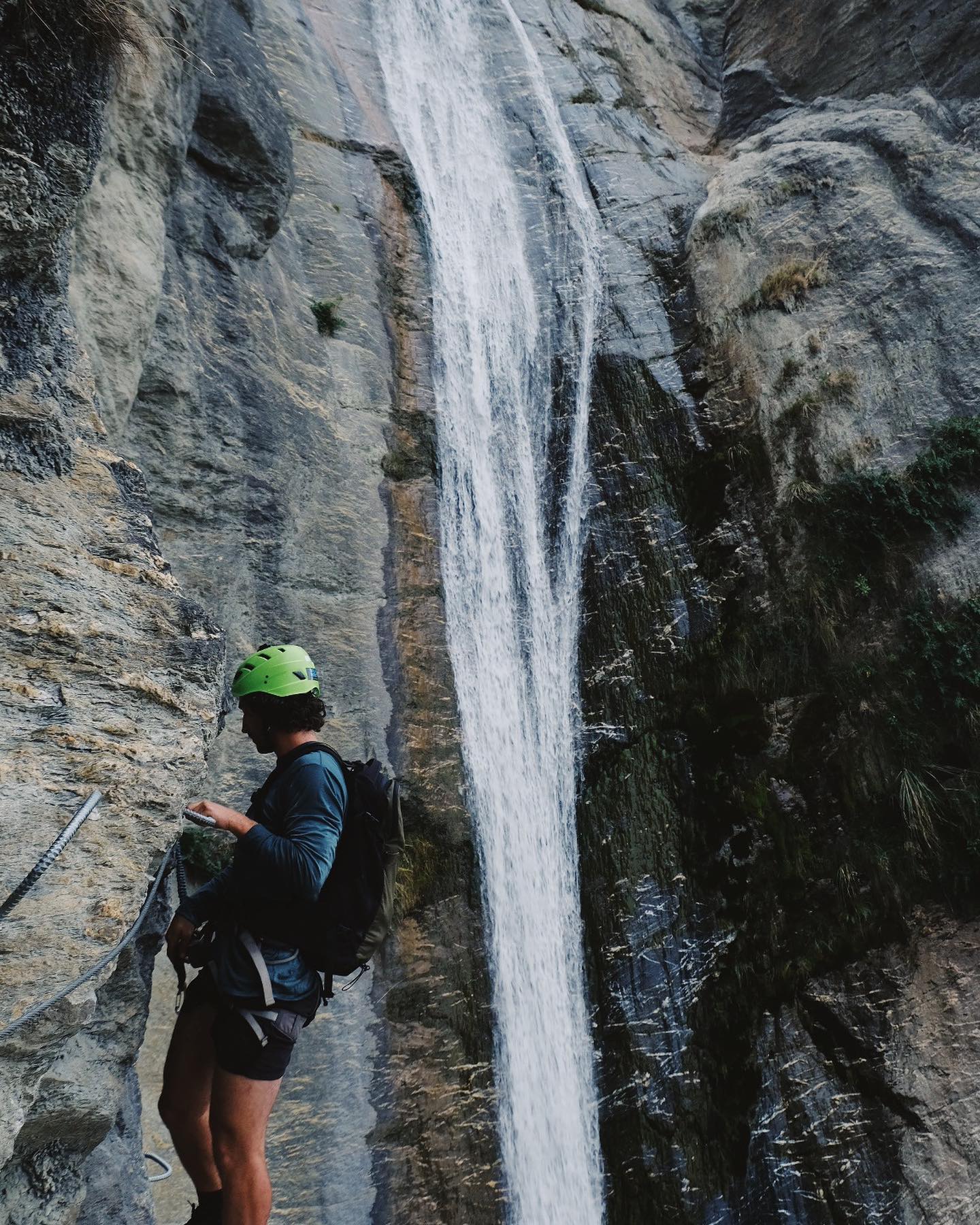 A climber in a green helmet standing next to a rockface with a waterfall in the background at Wildwire Wānaka New Zealand
